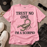 A pink graphic t-shirt laid flat on a gray concrete surface, surrounded by lifestyle props including a black baseball cap, black sunglasses, white sneakers, and dark denim jeans. The t-shirt features a black text graphic reading 'TRUST NO ONE' above a tan scorpion illustration, and 'I'M A SCORPIO' below.