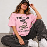 A woman sitting on a concrete floor wearing a pink graphic t-shirt that reads 'TRUST NO ONE I'M A SCORPIO' with a scorpion illustration, paired with dark gray washed jeans, white sneakers, and a black baseball cap.